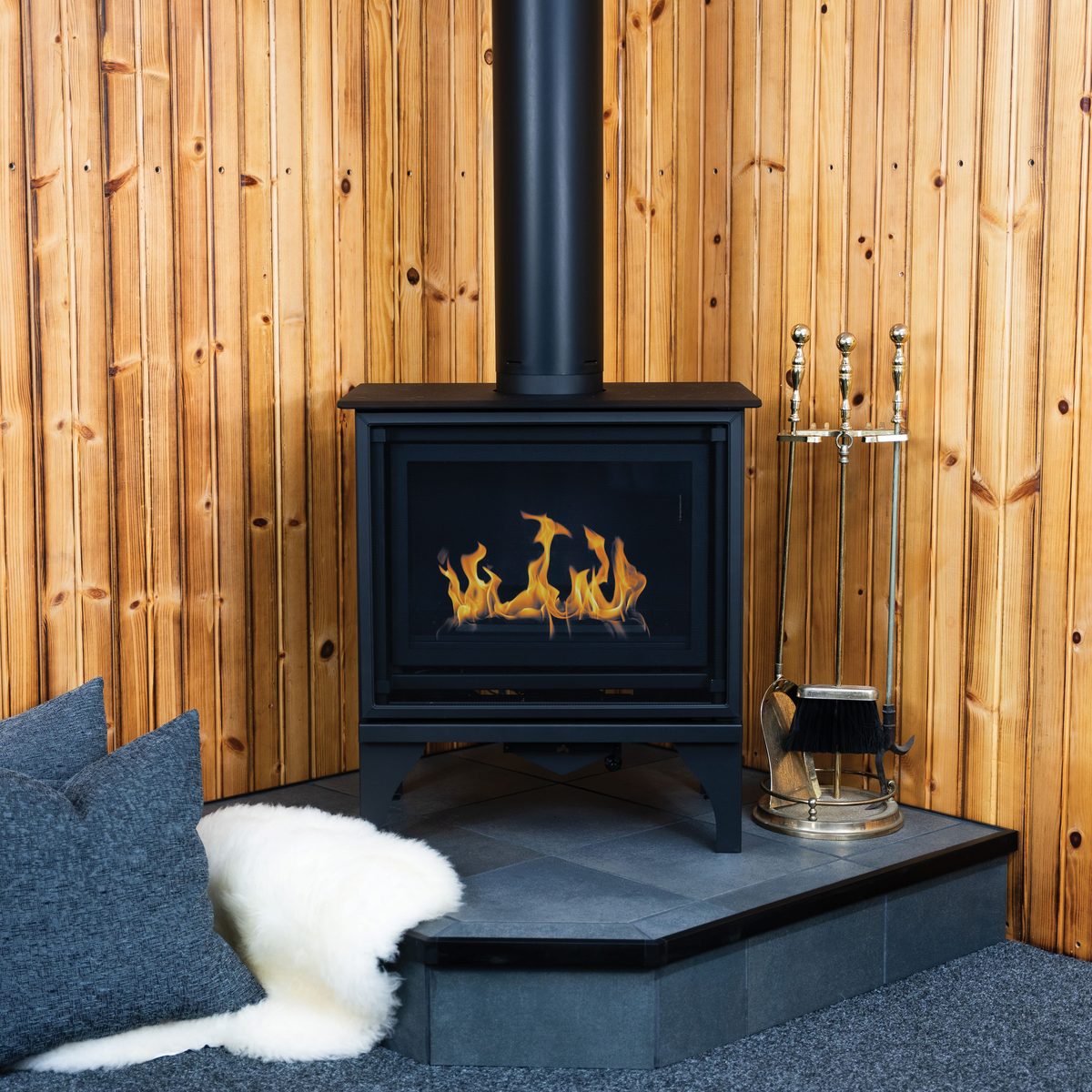 A wood stove and brass tool set on a Hearth Classics corner hearth pad against a knotty-pine wall